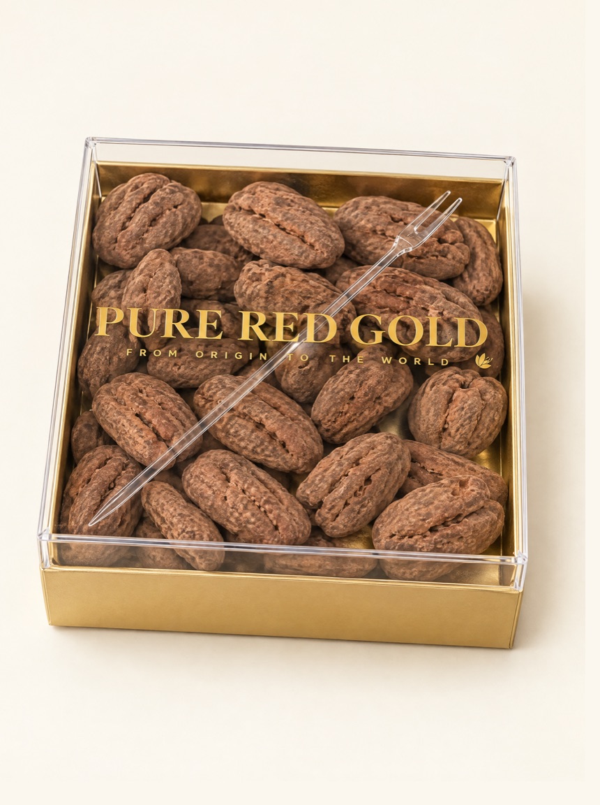 A square gold foil tray of cocoa-dusted pecan halves under a clear lift-off lid printed with the gold Pure Red Gold lockup, on warm ivory