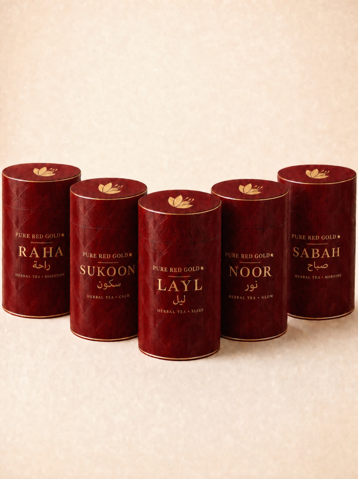 The Pure Red Gold herbal tea tubes — crimson cylinders with an embossed art-deco pattern and gold serif names, Layl in front with Sukoon and Noor behind, on warm ivory