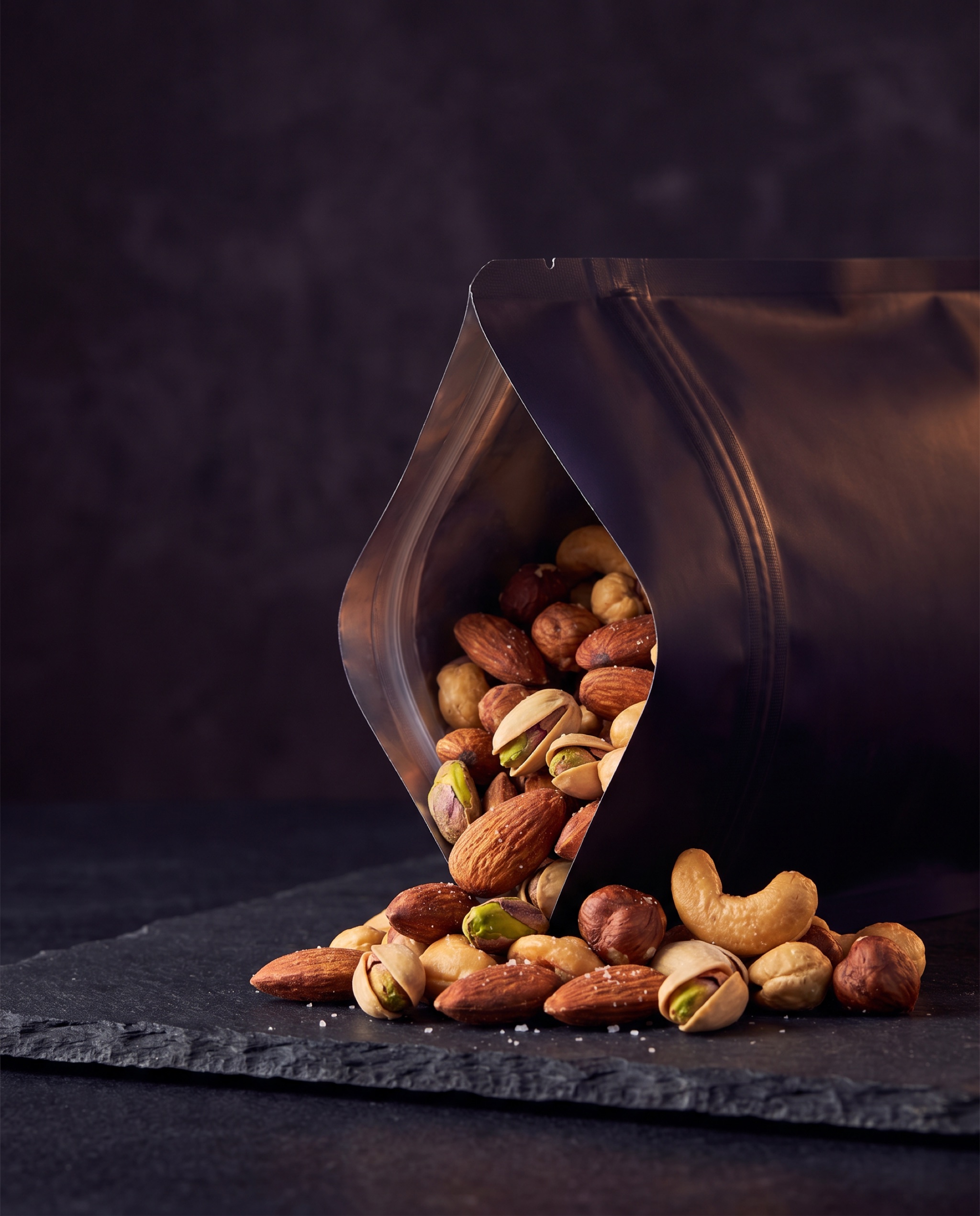 Roasted almonds, pistachios, cashews and hazelnuts spilling from a dark resealable pouch onto slate